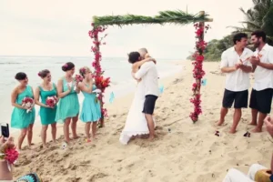 Casual beach wedding with the couple embracing under a floral arch surrounded by their wedding party