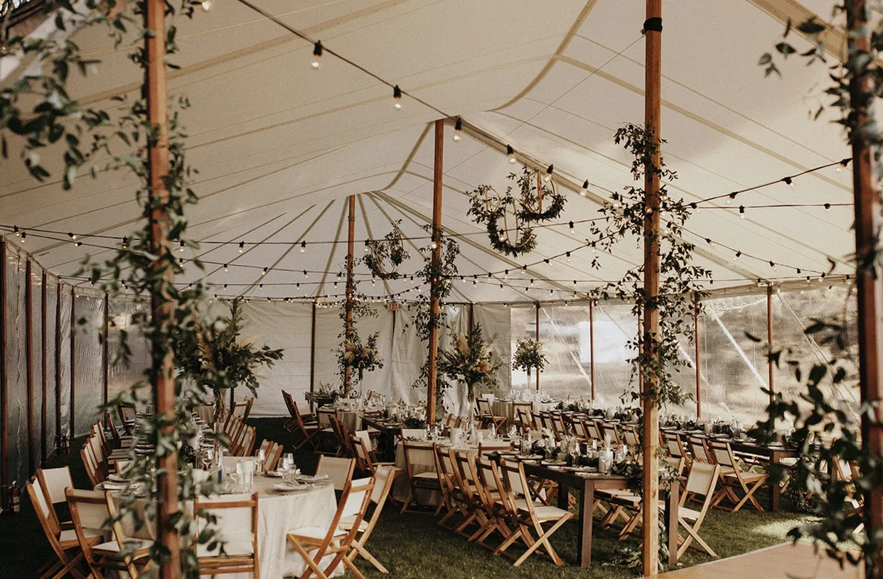 Rustic tent wedding reception with string lights, greenery and wooden chairs