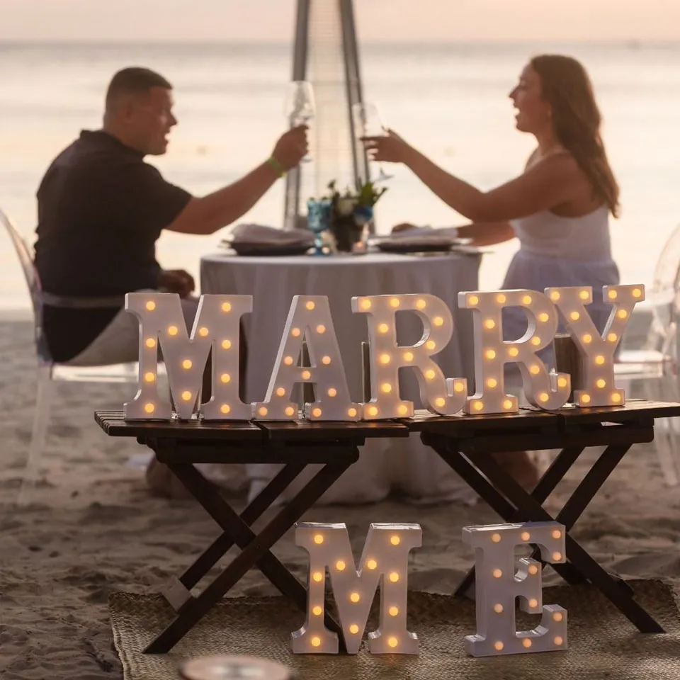 Couple toasting behind lit MARRY ME letters on West Bay Beach
