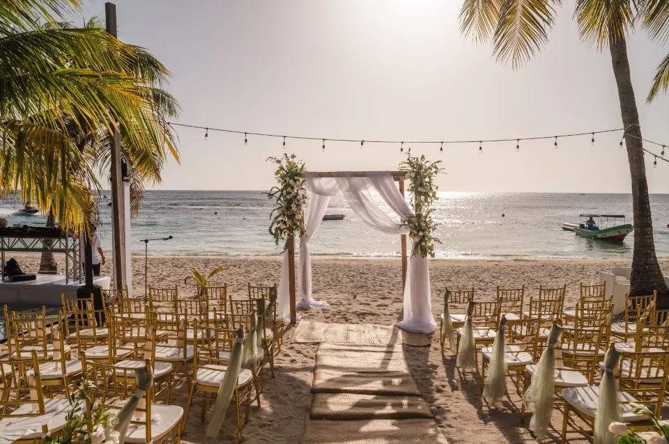 Beach ceremony setup with draped arch and gold chairs under palm trees