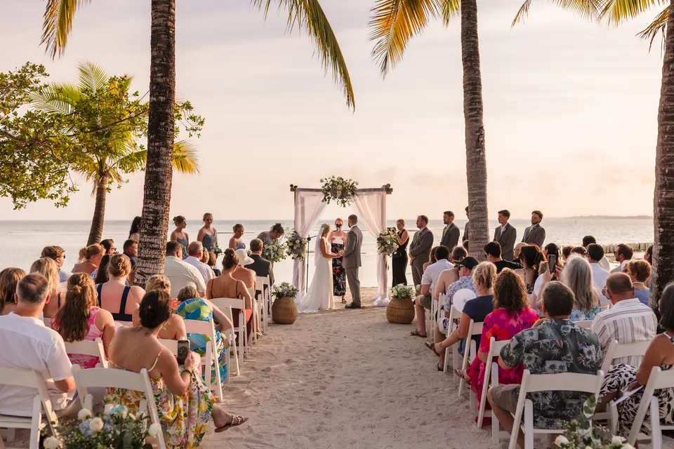 Guests watching Chloe and Avery exchange vows under a white draped arch at sunset at Bando Beach, Utila